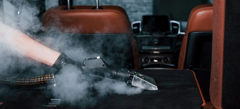 Worker using high-heat steam cleaner on light cloth seats inside a parked car